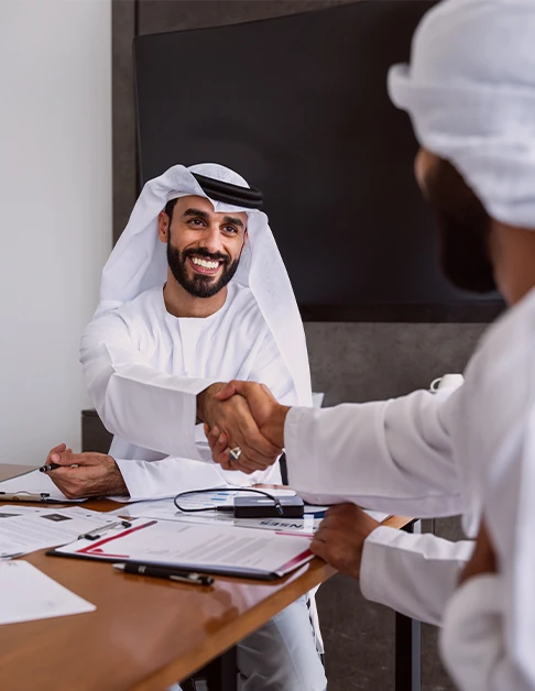 رجل مبتسم بالزي الوطني الأبيض يصافح رجلا آخر من فوق مكتب